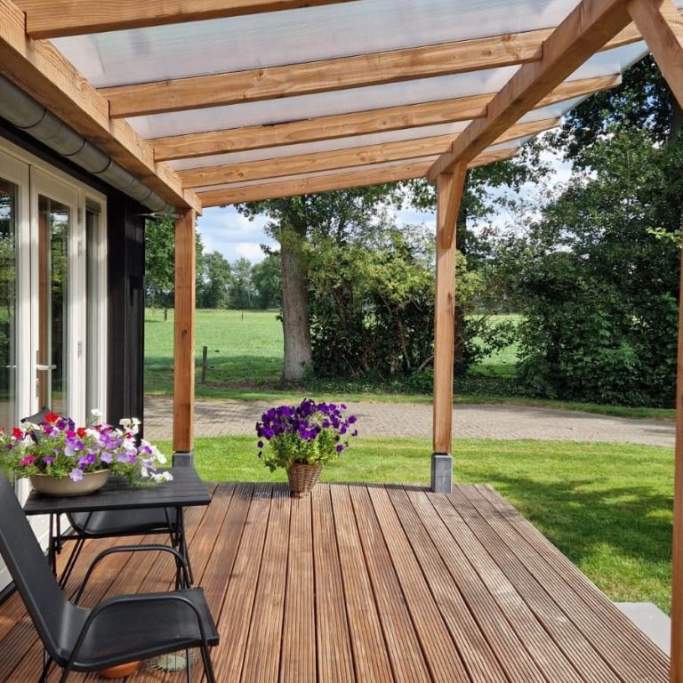 Overdekt terras met houten vloer, stoel en bloempotten in een groene omgeving.