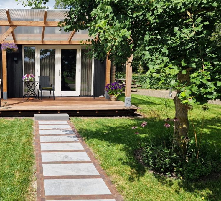 Een houten terras met paden en een huisje omringd door groen en bloemen.